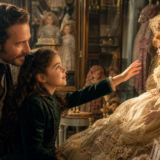 Young girl in a green velvet dress reaches toward an ornate porcelain doll in a Victorian-style doll shop, as an adult man smiles nearby among glass cabinets and dolls.