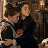 Two young girls hug warmly in a vintage living room by a lit fireplace, one holding a red heart-shaped cushion.