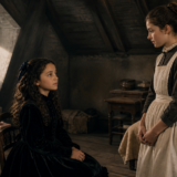 Two girls in period clothing converse in a dim attic; one sits in a black dress, the other stands in a gray dress with an apron, with a dollhouse and gifts nearby in the background.