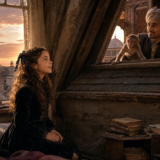 Young woman sits in a dusty attic at sunset, speaking with a man in a turban peering through a window, a small monkey beside him and books nearby.