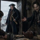 Three men in a weathered seaside tavern: a young man sits at a wooden table, an older man in a tricorn hat stands in the doorway with a brass spyglass, and another man on the right extends his hand.