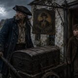 Pirate in a dark coat and tricorn hat pushes a wooden chest on a cart by the sea, beside a weathered inn sign reading Admiral Benbow Inn.