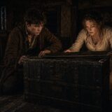Two adults crouch beside a large dark trunk in a dim, old room; a body lies on the floor nearby, lit by a candle.