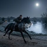 Man rides a galloping horse along a dirt path beside a frozen river under a bright full moon in a moonlit countryside.