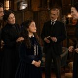Young girl in a blue velvet dress and bow stands among a somber-looking family in a richly furnished Victorian parlor.